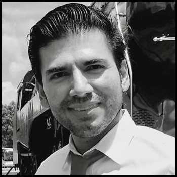 Headshot of Elver Ariza-Silva. Behind him is a bus. The photo is monochrome.