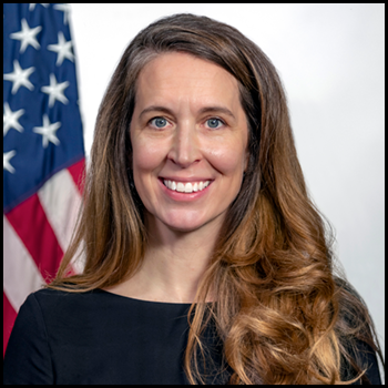 Headshot of Katy Kale. Behind her is a white backdrop with an American flag. She is wearing a black blouse.